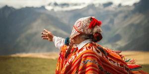 Indigenous Peruvian man wearing a traditional colorful Andean poncho, pointing towards the mountains.